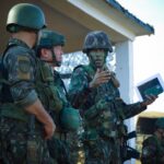 Group of soldiers in camouflage attending a briefing outdoors with tactical gear and helmets.