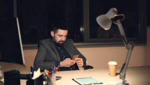 A businessman in a suit using a smartphone at his office desk at night.