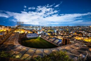 Aerial view of a brightly lit stadium surrounded by city buildings at dusk under a clear sky.
