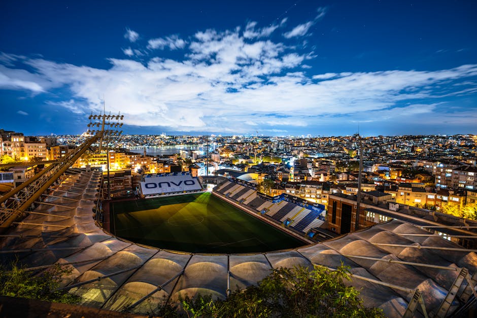Aerial view of a brightly lit stadium surrounded by city buildings at dusk under a clear sky.