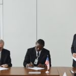 Executives signing international agreement with EU and US flags displayed on a wooden table.