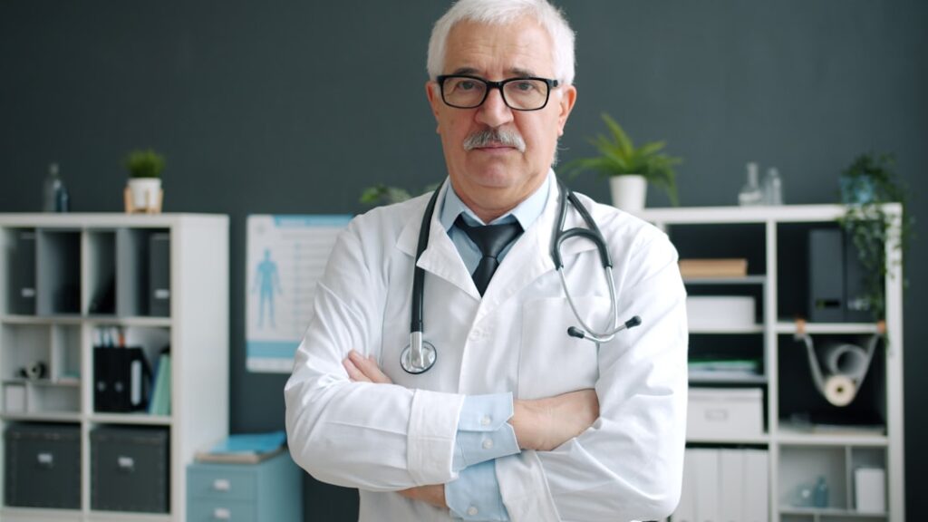 Portrait of confident experienced doctor in white gown and glasses standing in office room with arms crossed and looking