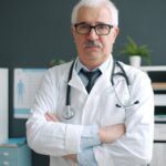 Portrait of confident experienced doctor in white gown and glasses standing in office room with arms crossed and looking