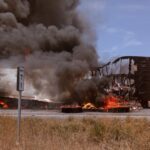A large cloud of smoke emerges from a burning cargo box beside the road.