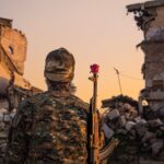 A soldier in camouflage with a red rose on rifle amidst ruined buildings at sunset.