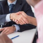 Two businessmen shaking hands across table, symbolizing agreement and partnership in an office environment.