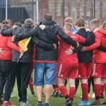 A soccer team huddles together on the field wearing red jerseys, showing unity and teamwork.